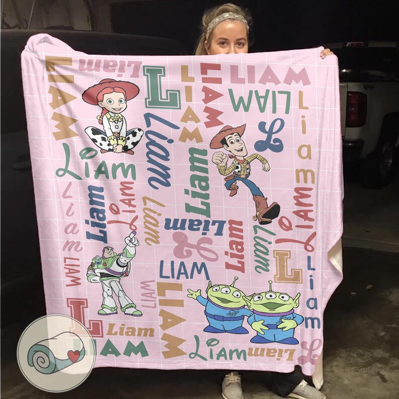 May include: A pink blanket featuring characters from the Toy Story franchise, including Jessie, Woody, Buzz Lightyear, and the Little Green Men. The name "Liam" is printed in various fonts and colors across the blanket.
