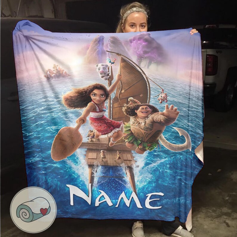 Może przedstawiać: Duży, niebieski koc z postaciami z animowanego filmu Moana. Koc ma żywą ilustrację Moany, Mauiego i innych postaci na łodzi, ze słowem "NAME" na dole. Koc jest trzymany przez osobę.