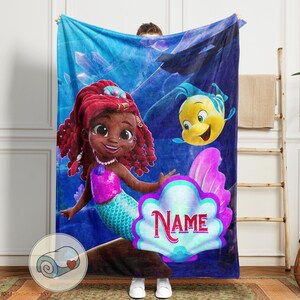 Peut inclure: Une couverture bleue avec une sir&egrave;ne de dessin anim&eacute; aux cheveux roux, un haut rose et une queue turquoise. Un poisson jaune nage &agrave; proximit&eacute;. Une grande coquille avec le mot "Name" est au centre. La couverture est tenue par une personne.