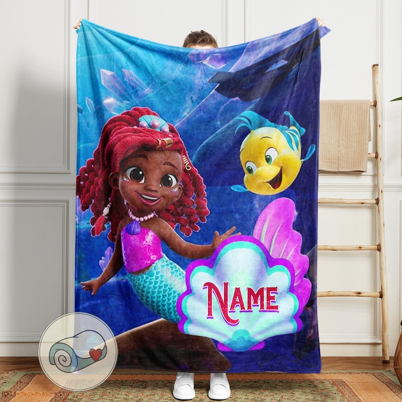 Peut inclure: Une couverture bleue avec une sir&egrave;ne de dessin anim&eacute; aux cheveux roux, un haut rose et une queue turquoise. Un poisson jaune nage &agrave; proximit&eacute;. Une grande coquille avec le mot "Name" est au centre. La couverture est tenue par une personne.