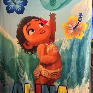 Pu&ograve; includere: Una coperta colorata con un'illustrazione a cartoni animati di una bambina che si allunga verso l'alto. Il disegno comprende onde oceaniche, fiori e il nome "ALINA" in basso. La bambina indossa una gonna floreale e un fiore rosso tra i capelli.