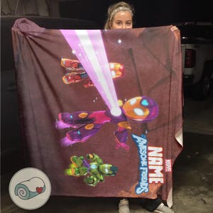Op de afbeelding: Een bruine deken met een kleurrijke cartoonillustratie van een superheld in een paars en roze pak, die een roze laserstraal afvuurt. De deken heeft de woorden "Awesome Friends" en "Name" en het Marvel-logo.