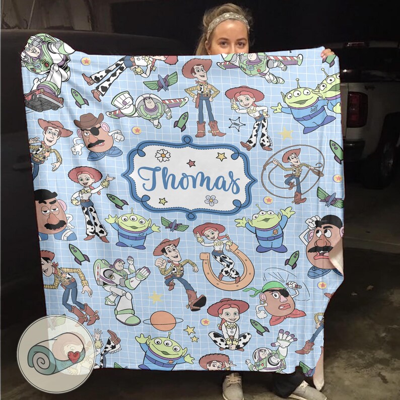May include: A light blue blanket featuring cartoon characters from the Toy Story franchise. The blanket has a grid pattern and includes the name "Thomas" in a decorative frame. Various characters are depicted in a repeating pattern.