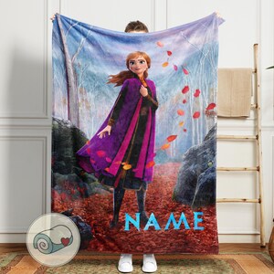 Pode incluir: Um cobertor macio com um personagem de um filme de anima&ccedil;&atilde;o popular, ambientado em um cen&aacute;rio de floresta com folhas caindo. O personagem est&aacute; vestindo uma roupa roxa e preta. A palavra "NAME" est&aacute; impressa em azul.