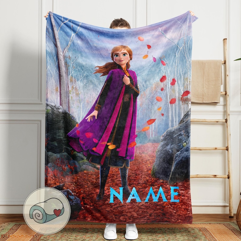 Pode incluir: Um cobertor macio com um personagem de um filme de anima&ccedil;&atilde;o popular, ambientado em um cen&aacute;rio de floresta com folhas caindo. O personagem est&aacute; vestindo uma roupa roxa e preta. A palavra "NAME" est&aacute; impressa em azul.