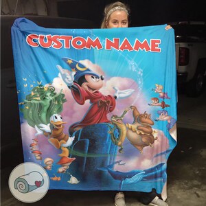 May include: A blue blanket featuring a colorful illustration of Mickey Mouse as a wizard, with other Disney characters. The blanket has the text "CUSTOM NAME" in red letters at the top. The blanket is held up by a person.