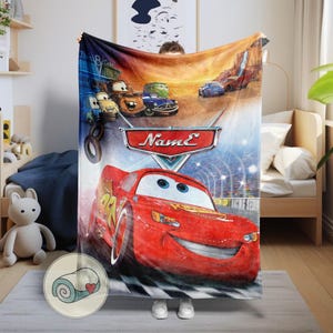 May include: A colorful blanket featuring characters from the animated movie "Cars." The design includes a red race car with the number 95, other cars, and a racing track. The blanket is held up by a person.
