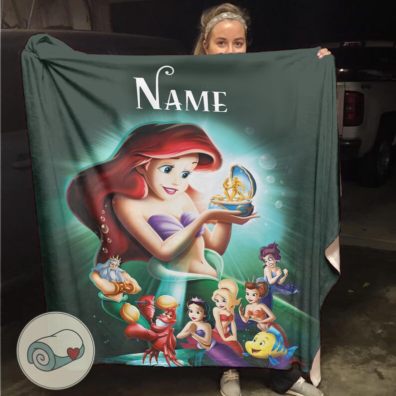 May include: A teal blanket featuring Ariel from The Little Mermaid, holding a treasure. The blanket has the word "Name" in white script at the top. Other characters from the movie are also featured on the blanket.