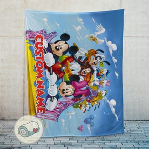 May include: A large, soft blanket featuring a colorful illustration of Mickey Mouse, Minnie Mouse, Donald Duck, Goofy, and other Disney characters. The blanket has a blue sky background with clouds and the text "CUSTOM NAME" in vertical letters.