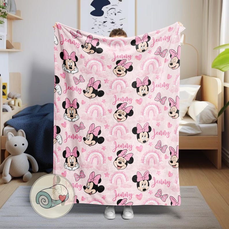 May include: Pink blanket featuring a repeating pattern of Minnie Mouse, rainbows, hearts, and butterflies. The name "Jenny" is written in a script font throughout the design. The blanket is a soft, cozy item.