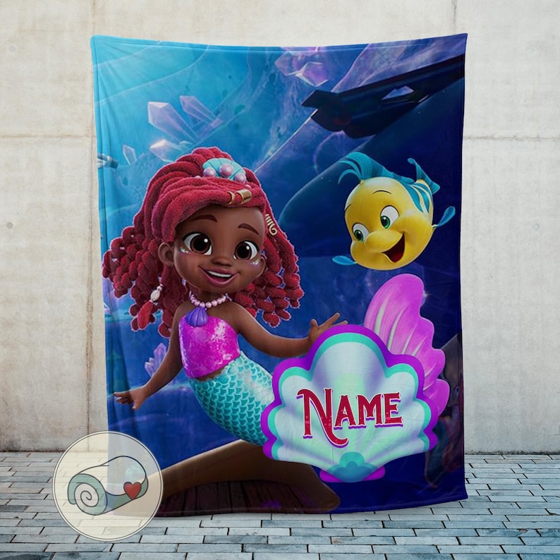 Peut inclure: Une couverture en peluche repr&eacute;sentant une sir&egrave;ne de dessin anim&eacute; aux cheveux roux, un haut rose et une queue turquoise. Un poisson jaune et bleu nage &agrave; proximit&eacute;. La couverture a un fond sous-marin bleu avec le mot "NAME" sur un coquillage.