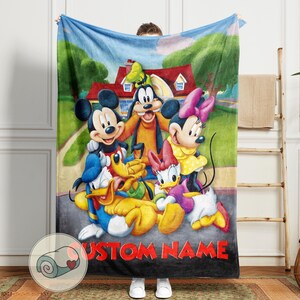 K&ouml;nnte beinhalten: Eine weiche Decke mit einer farbenfrohen Illustration von Mickey Mouse, Minnie Mouse, Goofy, Donald Duck und Daisy Duck vor einem Haus. Der Text "CUSTOM NAME" in Rot ist unten auf der Decke zu sehen.