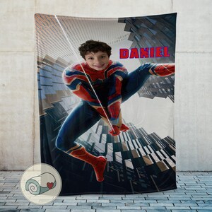 Op de afbeelding: Een gepersonaliseerde deken met een kind verkleed als Spider-Man, tegen een stadsgezicht. De naam "DANIEL" staat in rode letters. De deken is een zacht, knus item, perfect voor een kinderkamer of als cadeau.
