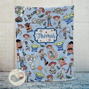 May include: A light blue blanket featuring various cartoon characters, including Woody, Buzz Lightyear, and Jessie. The blanket has a repeating pattern of the characters and space-themed elements. A central label reads "Thomas" in a decorative font.