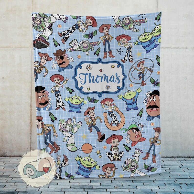 May include: A light blue blanket featuring various cartoon characters, including Woody, Buzz Lightyear, and Jessie. The blanket has a repeating pattern of the characters and space-themed elements. A central label reads "Thomas" in a decorative font.