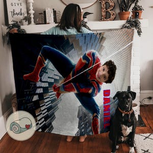 Op de afbeelding: Een gepersonaliseerde deken met een Spider-Man-ontwerp in rood, blauw en goud. De naam "DANIEL" is verticaal in het rood gedrukt. Een zwarte hond met een bruine halsband zit voor de deken.