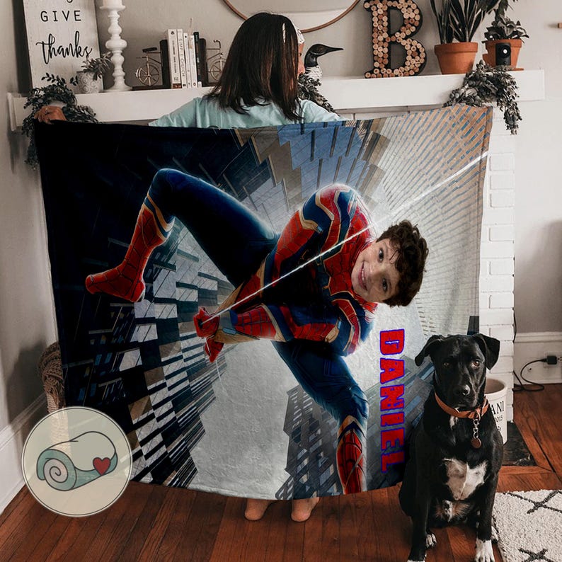 Op de afbeelding: Een gepersonaliseerde deken met een Spider-Man-ontwerp in rood, blauw en goud. De naam "DANIEL" is verticaal in het rood gedrukt. Een zwarte hond met een bruine halsband zit voor de deken.