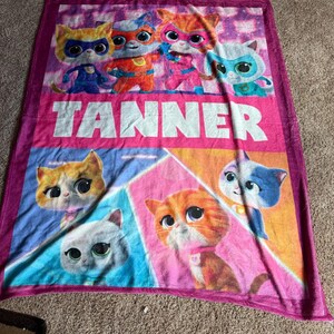 Pu&ograve; includere: Una coperta in pile rosa e viola con personaggi di gatti dei cartoni animati. La coperta ha il nome "TANNER" a grandi lettere bianche. La parte superiore mostra i gatti in costume da supereroi, mentre la parte inferiore presenta immagini ravvicinate dei gatti.