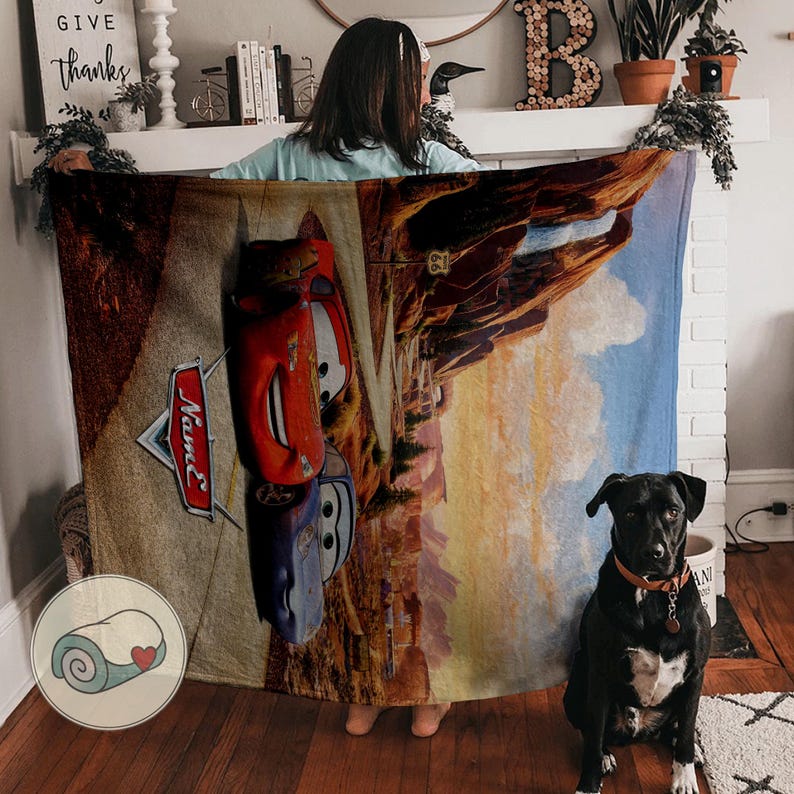 May include: A large, rectangular blanket featuring a design with the red car Lightning McQueen and the blue car, Mater, from the movie Cars. The blanket has a desert landscape background with a blue sky and a dog sitting in front of it.
