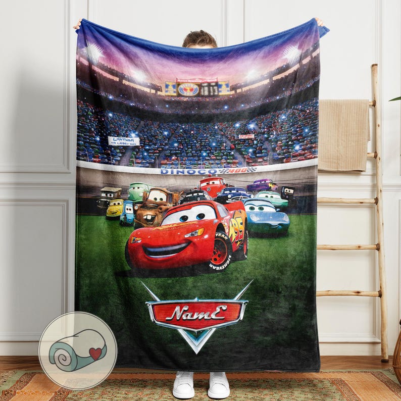 May include: A plush blanket featuring characters from the animated movie Cars. The blanket showcases Lightning McQueen and other race cars on a green field, with a stadium backdrop. The blanket has a red, blue, green, and black color scheme.