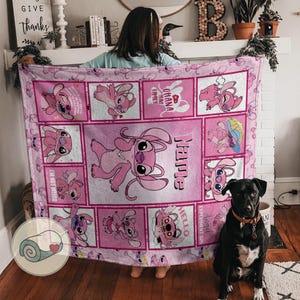 May include: Pink fleece blanket featuring a cartoon character in various poses within square frames. The blanket has the word "Name" and the phrase "Ohana means family" printed on it. A black dog sits in front of the blanket.