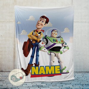 Pu&ograve; includere: Una morbida coperta con Woody e Buzz Lightyear di Toy Story. Woody indossa una camicia gialla, jeans blu e un cappello da cowboy. Buzz Lightyear &egrave; in una tuta spaziale bianca e verde. La coperta ha uno sfondo di cielo blu e la parola "NAME".