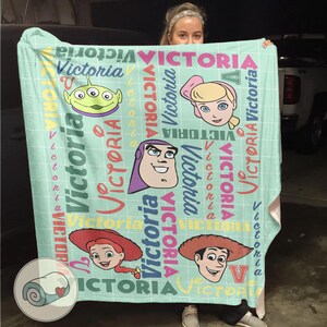 K&ouml;nnte beinhalten: Eine hellt&uuml;rkisfarbene Decke mit Cartoon-Charakteren und dem Namen "Victoria" in verschiedenen Farben. Die Decke hat ein Gittermuster und enth&auml;lt Bilder eines Aliens, Bo Peep, Buzz Lightyear, Jessie und Woody.