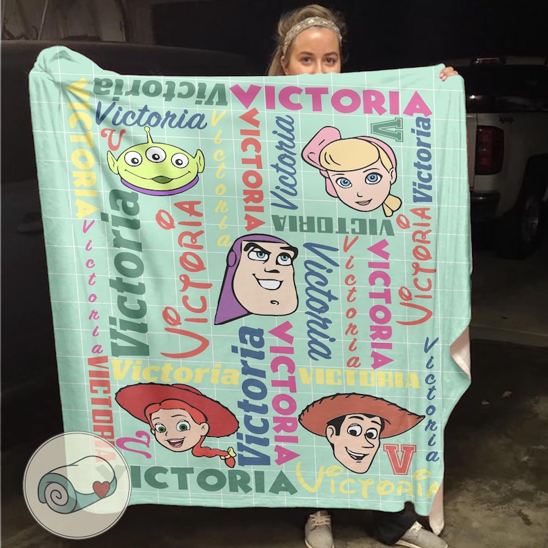 K&ouml;nnte beinhalten: Eine hellt&uuml;rkisfarbene Decke mit Cartoon-Charakteren und dem Namen "Victoria" in verschiedenen Farben. Die Decke hat ein Gittermuster und enth&auml;lt Bilder eines Aliens, Bo Peep, Buzz Lightyear, Jessie und Woody.