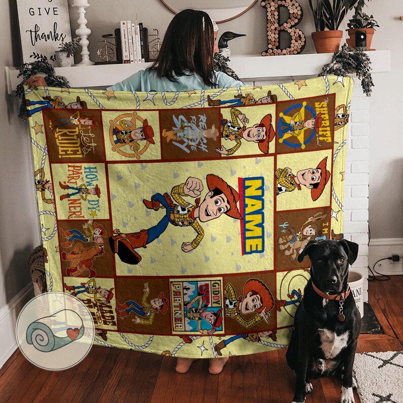 May include: A yellow blanket featuring a cartoon cowboy character in various poses, with the word "NAME" in a vertical blue and yellow box. The blanket has a brown border and includes the words "SHERIFF", "GIDDY PARTNER", and "HOWDY PARTNER". A black dog sits in front of the blanket.