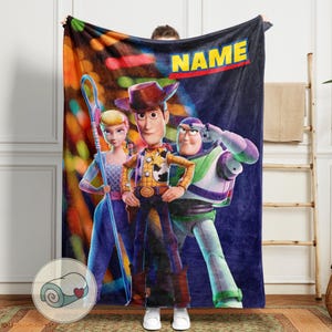 Puede incluir: Una manta de felpa con personajes de la pel&iacute;cula animada Toy Story. La manta muestra a Woody, Buzz Lightyear y Bo Peep sobre un fondo azul oscuro. La palabra "NAME" se muestra en amarillo.