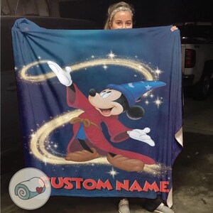 Op de afbeelding: Een blauwe deken met Mickey Mouse als de Tovenaarsleerling uit Fantasia. Mickey draagt een rode mantel en een blauwe tovenaarsmuts, met een wervelend goudkleurig ontwerp en de tekst "CUSTOM NAME" onderaan.