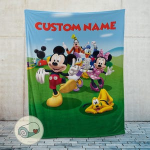 Pu&ograve; includere: Una coperta in pile blu con Topolino, Minnie, Paperino, Pippo e Pluto su un campo verde con uno sfondo di cielo blu. Le parole "CUSTOM NAME" sono stampate in rosso in alto.