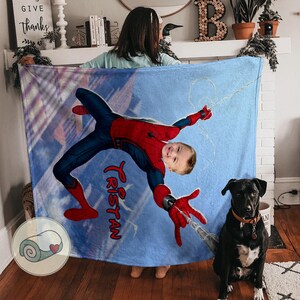 Op de afbeelding: Een blauwe deken met een Spider-Man-ontwerp, gepersonaliseerd met de naam "Tristan" in rode letters. Het ontwerp toont het gezicht van een kind op het lichaam van de superheld. Een zwarte hond is zichtbaar op de voorgrond.