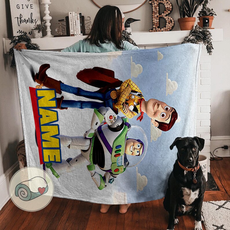 Pu&ograve; includere: Una coperta azzurra con Woody e Buzz Lightyear di Toy Story contro un cielo azzurro con nuvole bianche. La coperta ha la scritta "NAME" in giallo e rosso. Un cane nero &egrave; seduto davanti alla coperta.