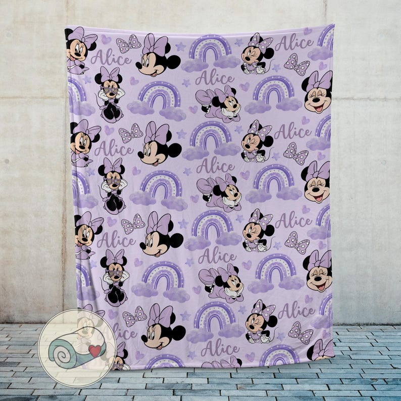 Op de afbeelding: Een lavendelkleurige deken met een herhalend patroon van Minnie Mouse, strikken, regenbogen, wolken en sterren. De naam "Alice" staat in het hele ontwerp geschreven. De deken is gemaakt van zacht materiaal.