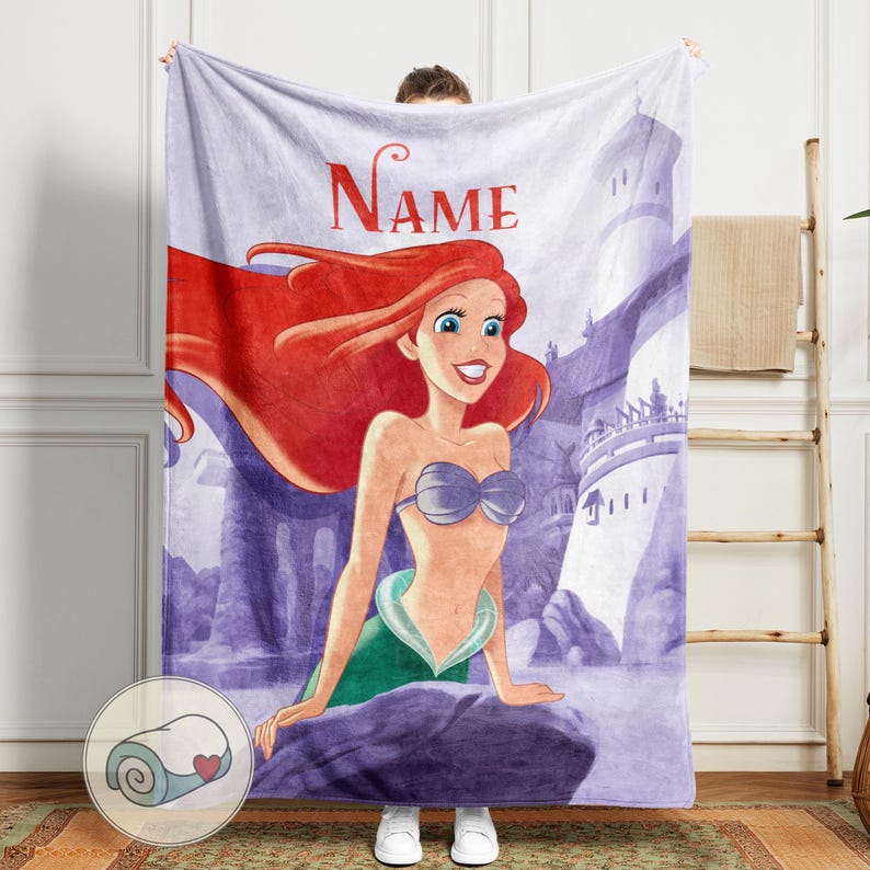 May include: A plush blanket featuring a cartoon illustration of a mermaid with red hair, a blue seashell top, and a green tail. The blanket has the word "Name" in red script at the top, with a purple castle background.