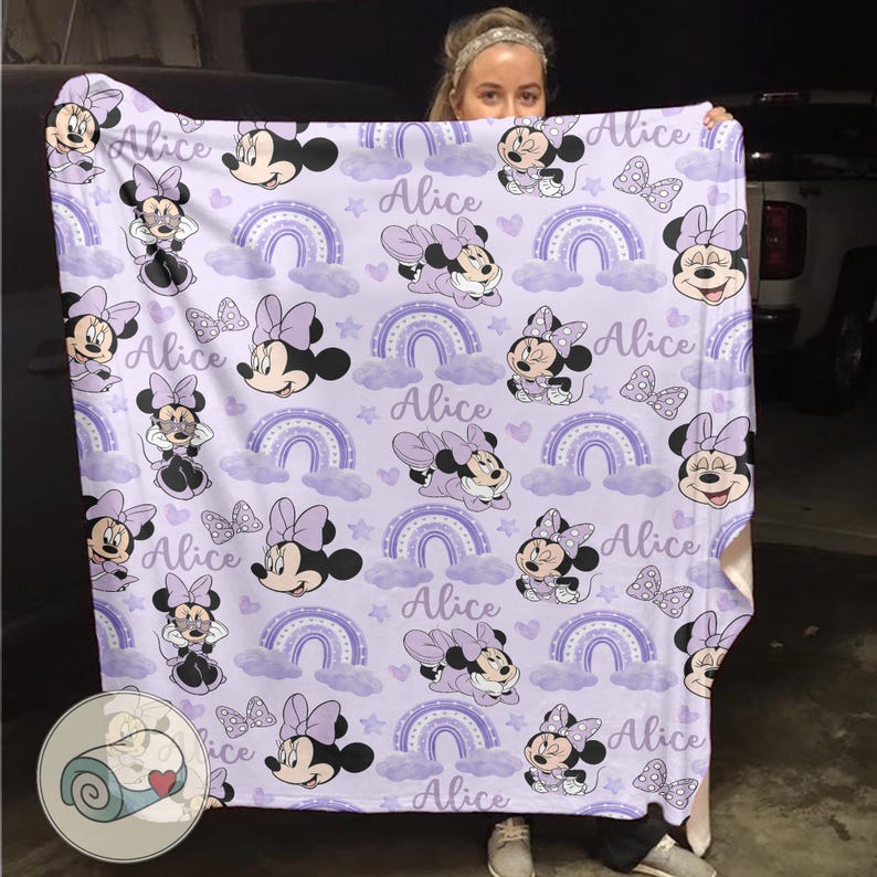Op de afbeelding: Een lavendelkleurige deken met een herhalend patroon van Minnie Mouse, regenbogen, sterren en strikken. De naam "Alice" staat in het hele ontwerp geschreven. De deken is een zacht, gezellig item.