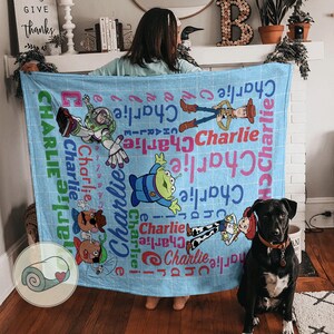 May include: A light blue blanket featuring characters from the Toy Story movie, including Buzz Lightyear, Woody, and an alien. The name "Charlie" is printed in various colors and fonts across the blanket. A black dog is sitting in front of the blanket.