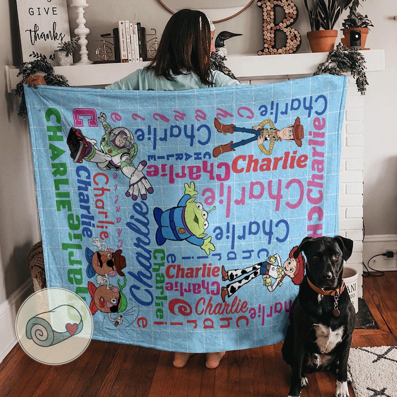 May include: A light blue blanket featuring characters from the Toy Story movie, including Buzz Lightyear, Woody, and an alien. The name "Charlie" is printed in various colors and fonts across the blanket. A black dog is sitting in front of the blanket.