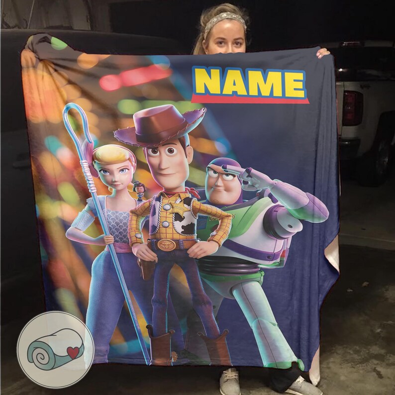 Puede incluir: Una manta rectangular grande con personajes de la pel&iacute;cula animada "Toy Story". La manta muestra a Woody, Buzz Lightyear y Bo Peep sobre un fondo azul oscuro. La palabra "NAME" est&aacute; impresa en amarillo. Un peque&ntilde;o logotipo circular est&aacute; en la esquina inferior izquierda.