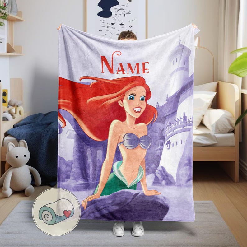 May include: A plush blanket featuring a cartoon illustration of a mermaid with red hair, a silver seashell bra, and a green tail. The blanket has the word "Name" in red script at the top. The background includes a castle and purple rocks.