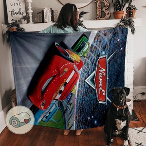 May include: A large, rectangular blanket featuring a vibrant design of a red race car, other colorful cars, and a racing scene. The blanket has a blue background with the word "Name" on a red sign.