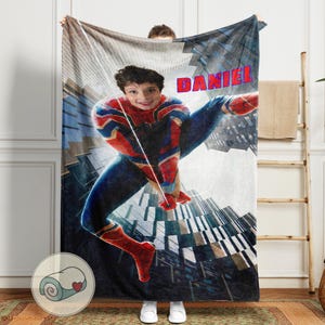 Op de afbeelding: Een gepersonaliseerde deken met een jongeman verkleed als Spider-Man, met de naam "DANIEL" in rode letters. Het ontwerp bevat een stadsgezicht achtergrond en is overwegend blauw, rood en grijs.