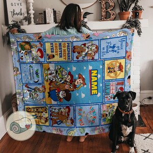 May include: A light blue blanket with Toy Story characters. The blanket has a central panel with the word "NAME" and various character illustrations. Other panels include phrases like "You've got a Friend in Me" and "To Infinity and Beyond."