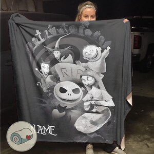 May include: A dark gray blanket featuring characters from The Nightmare Before Christmas. The design includes Jack Skellington, Sally, and other characters in a grayscale illustration. The blanket has a personalized name at the bottom.
