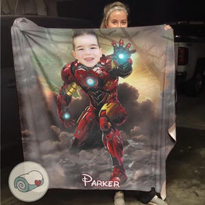 May include: A personalized blanket featuring a child's face superimposed on an Iron Man illustration. The blanket is predominantly red and gold, with a background of clouds and a sunset. The name "Parker" is printed on the blanket.