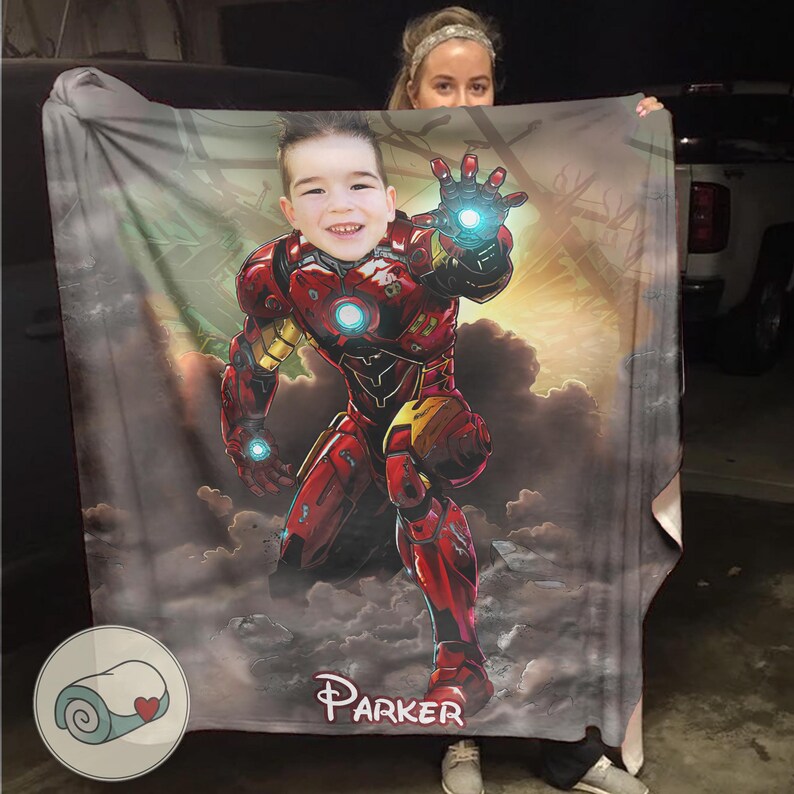 May include: A personalized blanket featuring a child's face superimposed on an Iron Man illustration. The blanket is predominantly red and gold, with a background of clouds and a sunset. The name "Parker" is printed on the blanket.
