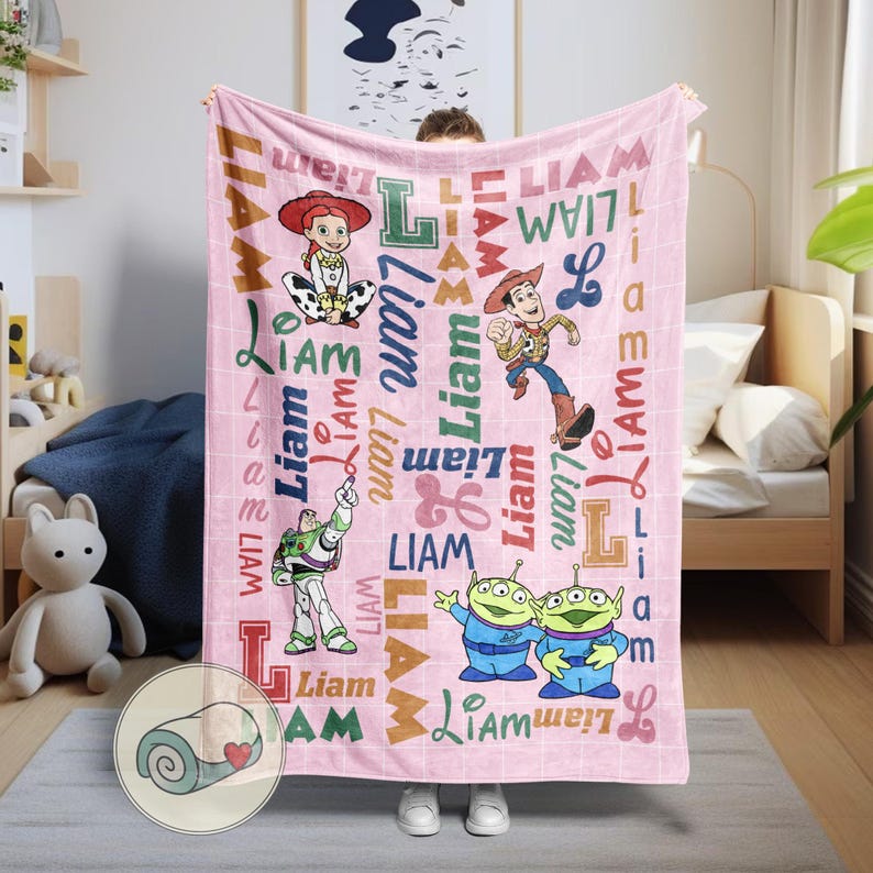 May include: A pink blanket featuring characters from the animated film "Toy Story." The blanket has the name "Liam" printed in various fonts and colors. The blanket features images of Woody, Buzz Lightyear, Jessie, and the aliens.