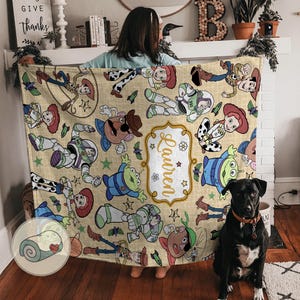 May include: A beige blanket featuring cartoon characters from a popular animated film. The blanket has a repeating pattern of characters, including a cowboy, space ranger, and cowgirl. The name "Lauren" is displayed in a decorative frame.