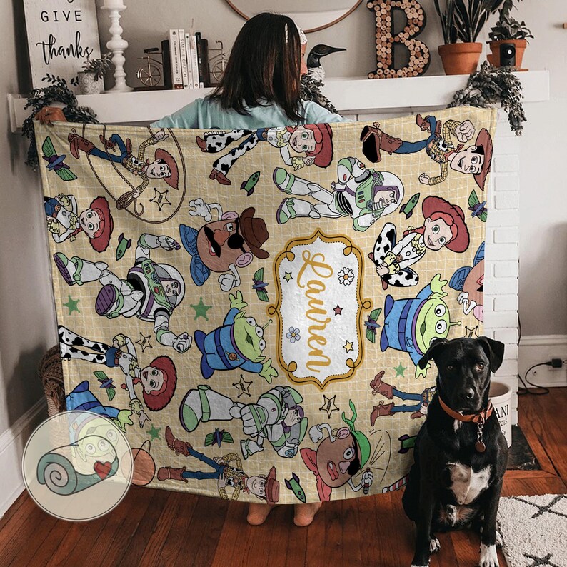 May include: A beige blanket featuring cartoon characters from a popular animated film. The blanket has a repeating pattern of characters, including a cowboy, space ranger, and cowgirl. The name "Lauren" is displayed in a decorative frame.